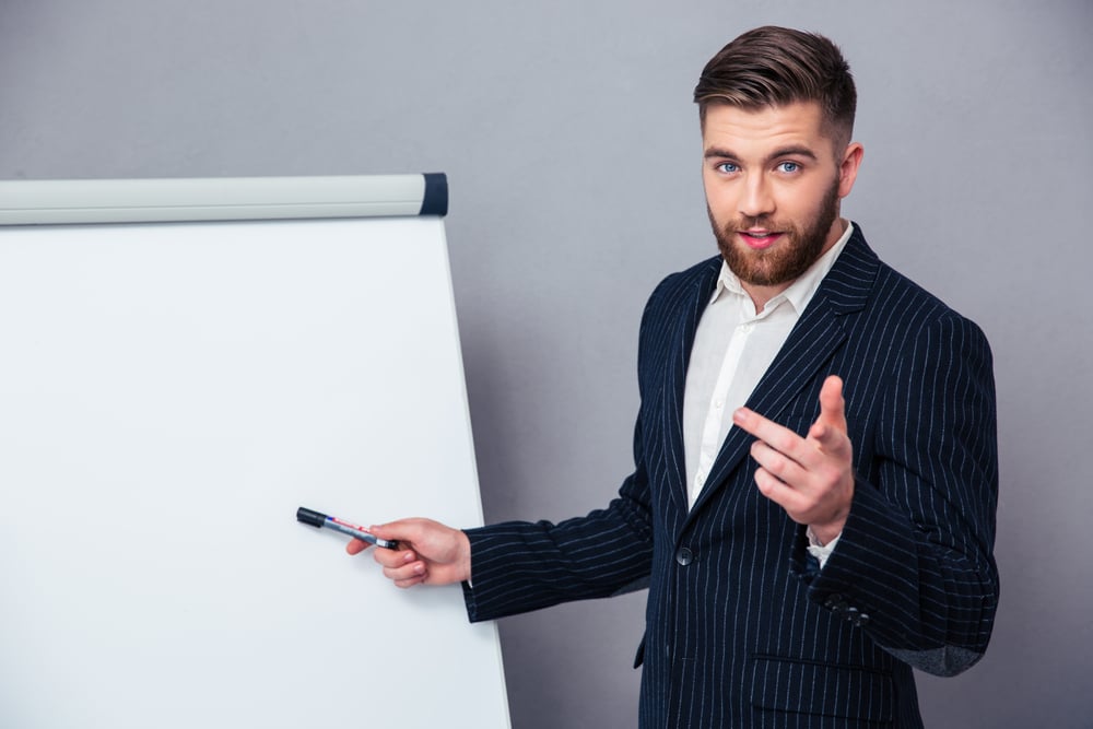 Portrait of a young businessman in suit presenting something on blank board over gray background